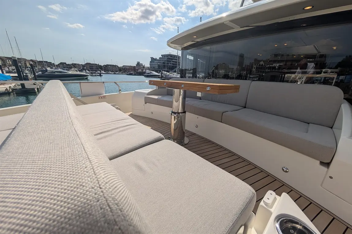 A modern boat deck featuring grey upholstered seating, a wooden table, and views of the marina under a clear blue sky.